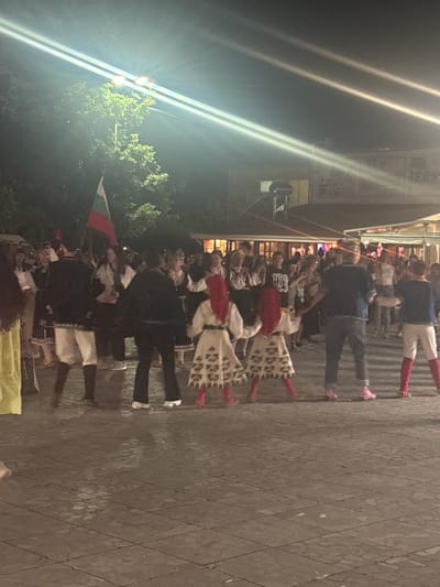 The image depicts an outdoor gathering at night, likely a cultural event or festival, in the city of Parvomai, Bulgaria. Several people are visible, some dressed in traditional Bulgarian folk costumes, including embroidered dresses and headwear. A Bulgarian flag is prominently displayed, held by someone in the crowd. The scene is illuminated by artificial lights, creating strong light beams across the frame. In the background, there is a building with some lighting, suggesting it might be a venue or establishment. The ground appears to be paved with stone tiles. Some individuals are engaged in dancing or observing the activities. The overall atmosphere suggests a lively community event.