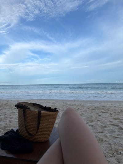 A first-person perspective shot captures a tranquil beach scene in Ao Cho, Thailand. In the foreground, a person's bare legs and knee are visible, suggesting they are reclining on a lounge chair. To the left, a woven straw beach bag with dark pom-poms and straps sits on a dark wooden surface. A crumpled black garment is placed next to the bag. The sandy beach stretches out to the ocean, where gentle waves roll onto the shore under a partly cloudy blue sky.