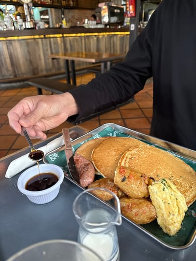 A person in a black long-sleeved shirt is holding a silver tray with pancakes, potatoes, and an omelet. A knife and fork are also on the tray. The person is using a spoon to add syrup from a small white container onto the food. A glass pitcher with milk sits beside the tray. The scene takes place at a restaurant in the city of La Tijera, Mexico. The restaurant has a wooden staircase and appears to have various items displayed on shelves.