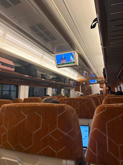 This media file captures the interior of a train carriage, likely in Dodoma, Tanzania, given the location context. The scene is oriented vertically, suggesting the photo was taken with the camera rotated. The train carriage features rows of orange seats with a geometric pattern, indicating a passenger seating area. Some seats are occupied by the heads and shoulders of people, though their faces are not clearly visible. A laptop is open on one of the seats, its screen displaying content.
Along the left side of the carriage, a long panel runs the length of the wall, featuring air vents and a small screen displaying a news broadcast with a flag and a speaker. The screen shows text, part of which appears to read "EST: DAR ES SALAAM". Luggage is visible in the overhead compartments and on the floor, including a pink suitcase. The lighting appears to be artificial, suggesting the time of day is not strongly indicated, though the ambiance is relatively dim. The overall impression is of a typical journey on a passenger train, with individuals engaged in their own activities or resting during transit. The visible text "EST: DAR ES SALAAM" provides a potential location cue, referring to Dar es Salaam, a major city in Tanzania, suggesting the train might be traveling to or from this location.