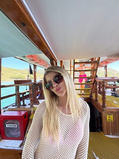 An eye-level indoor shot on what appears to be a boat or ferry in Menderes, Turkey, captures a blonde woman in sunglasses taking a selfie or being photographed. The image is rotated 90 degrees clockwise.

The main subject, positioned on the right side of the frame, is a woman with long, straight blonde hair, wearing large sunglasses and a light-colored, open-knit mesh top over a pink inner garment. She pouts playfully at the camera. Her right shoulder and chest are visible, with a small, round pendant on a delicate chain around her neck.

The setting is a wooden-paneled interior of a vessel, with prominent wooden railings and furniture structures. Through the boat's side, a bright, sunny daytime scene unfolds: clear blue sky, turquoise water, and a lush, green, hilly coastline in the distance. Another person, partially visible in the mid-ground, wears a white top and pink shorts, seemingly navigating or resting near a set of wooden stairs.

Notable details include a yellow "CAUTION WET FLOOR" sign attached to a wooden beam in the upper center, and a red box labeled "YANGIN" (Turkish for "FIRE") in the lower right, likely a fire extinguisher cabinet. The overall atmosphere suggests a leisurely boat trip on a beautiful, sunny day.