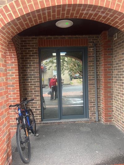 The image depicts a brick-lined entrance or archway in Mole Valley, United Kingdom. Within the archway is a modern gray-framed window reflecting a street scene. A person in a red jacket is visible in the window reflection. A blue and orange mountain bike lies on the gray asphalt outside the entrance, partially obscuring the view of the street. On the left wall of the archway, there is a white circular device with a green light. The brickwork is primarily red, with some areas featuring darker, almost brown bricks.