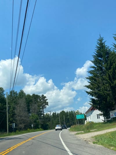 The image depicts a rural road in Saint-André-Avellin, Canada, on a sunny day with a blue sky and scattered clouds. A paved road with yellow double lines curves to the right. On the left side of the road, there are power lines strung across the frame and a dense line of evergreen trees. On the right side of the road, there is a small white house with a red roof, surrounded by grass and trees. A green road sign indicates directions to "CHENNEVILLE 10", "RIPON 8", and "MONTMELIANT 17". Two cars, a white SUV and a black sedan, are driving on the road, heading in the same direction as the viewer. The overall scene suggests a quiet, scenic drive through a wooded area.