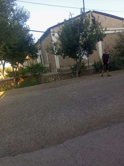A young boy stands on an asphalt road, looking to his right. He is wearing a black t-shirt and dark shorts. Behind him, an old, weathered building with a light-colored facade and brick accents stands under a clear sky. Trees with green foliage surround the building and the boy. The building appears to be a residential home, with windows and a damaged roofline. A low stone wall separates the building from the road. The sun is low in the sky, casting long shadows and suggesting it is either early morning or late afternoon. The setting appears to be a rural or semi-rural area, possibly in Tahmach, Uzbekistan, given the architectural style and vegetation. The overall mood is calm and still. No text is visible in the image.