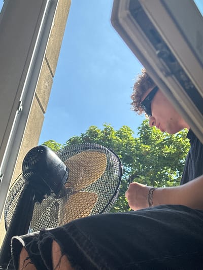 A low-angle, medium shot captures a young person with curly hair wearing black sunglasses and a dark t-shirt, sitting in front of a black oscillating fan. The fan's protective grille is visible, and the fan blades are a light yellow color. The person's arm, adorned with a silver bracelet, is bent and their fist is clenched. The background consists of lush green trees and a clear blue sky, suggesting an outdoor setting. The scene appears to be captured from an elevated position, possibly a window or balcony, with a concrete building element visible on the left side of the frame. The image composition, with the fan and person partially framing the view, suggests a focus on creating a comfortable, breezy atmosphere. This scene is likely taking place in Hamm, Germany, as implied by the context.