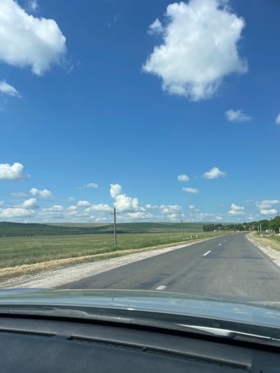 The image depicts a paved road in Coșernița, Moldova, stretching into the distance. The road is bordered by a grassy field on the left and a gravel shoulder on the right. Telephone poles with wires run alongside the road, indicating rural infrastructure. In the far distance, a small cluster of houses with green roofs can be seen, suggesting a village or hamlet. The sky is a vibrant blue with scattered white, fluffy clouds. The sunlight suggests it is daytime, likely mid-morning or afternoon. The overall scene conveys a sense of peacefulness and rural tranquility. There is no visible text in the image.