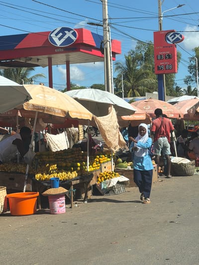 Here is a detailed description of the scene captured in the image:

**Location:** The image is taken in Pemba, Mozambique. This is evidenced by signs with Mozambican domain names like ".co.mz" and references to the mobile payment system "m-pesa", which is common in East Africa. The gas station chain "Exito Energia" is also visible, aiding in location identification.

**Environment:** It appears to be a bustling street scene. The ground is paved and constitutes a road or path for vehicles and pedestrians. The weather is sunny and clear.

**People:**
*   Several individuals are present, including vendors and a person wearing a blue dress and a headscarf, potentially a student or a local resident. There is a man in a red shirt standing beside the woman in the blue outfit.

**Objects:**
*   A prominent feature is an "Exito Energia" gas station with a price display sign indicating the prices for gasoline (83.57) and diesel (79.88).
*   Street vendors have set up stalls with umbrellas to shield their goods and customers from the sun.
*   Various fruits, including oranges and bananas, are displayed for sale. Other items such as buckets and other produce are also visible.
*   The umbrellas have advertisements for the mobile carrier "5G", a Super Movie App, and other promos.

**Actions/Interactions:**
*   People are engaged in commerce and walking along the street.
*   The woman in the blue dress is captured in what appears to be a moment of movement, perhaps walking or pausing by the vendors.

**Other Details:**
*   Power lines are visible overhead, indicating the presence of electrical infrastructure in the area.
*   Palm trees in the background suggest a tropical environment consistent with the location of Pemba in Mozambique.