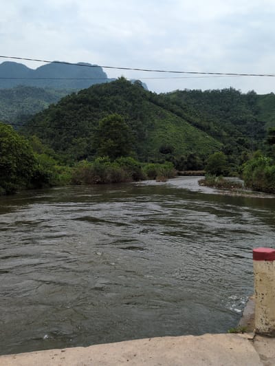 The image depicts a wide, murky river flowing through a verdant, mountainous landscape. The river occupies the foreground and midground, its surface rippled by a current. Lush green vegetation covers the rolling hills and mountains in the background, with trees and shrubs lining the riverbanks. The sky is overcast, contributing to a somewhat subdued lighting. In the lower right corner, a concrete structure with a red-capped post is partially visible, suggesting the presence of a bridge or embankment. The scene is serene and natural, typical of a rural setting in Vietnam. This specific location is identified as Ngọc Đường Commune, Vietnam.