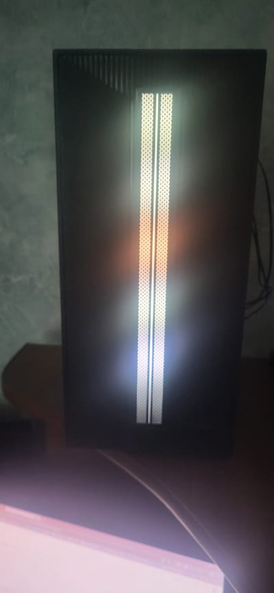 A close-up, slightly blurry, vertical shot shows the front panel of a black computer tower. The panel features two vertical, illuminated white grilles with a subtle orange glow emanating from the center. To the left of the grilles, the panel has a textured vertical stripe pattern. The background is a wall with a mottled, light green texture. A dark wooden surface, likely a desk or shelf, is visible at the bottom of the frame, and a bright, out-of-focus area at the bottom suggests a monitor or light source. The overall impression is of an indoor setting, possibly a home office or gaming room. The image does not contain any elements that would allow for its localization to the city of Гамово, Russia.