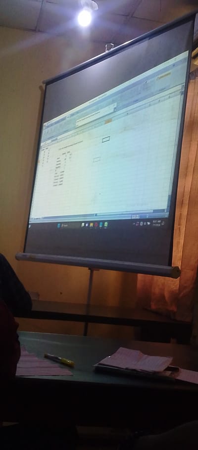 This media file depicts an indoor scene, likely a classroom or meeting room, in Achara Ndiagu Omegio, Nigeria. The primary focus is a large projection screen displaying a spreadsheet, resembling Microsoft Excel, with various data entries. The spreadsheet's content is partially visible, showing columns and rows with numerical and textual information. The top bar of the spreadsheet indicates a file name or title, though it is not fully legible.
In the foreground, a dark desk is visible, holding several papers and a yellow marker pen. Part of a person's arm and shoulder can be seen on the left, suggesting someone is seated at the desk. The setting appears to be during the day, as indicated by the overhead light and the general illumination, although the overall lighting is somewhat dim. The wall behind the projection screen is a light, textured color, and a curtain is visible on the right side of the frame.
The time displayed on the bottom right of the screen is 10:17 AM, with the date likely corresponding to the current day. The scene conveys a sense of a presentation or learning session in progress. There are no visible weather cues. The focus is on the educational or informational content being projected, with the surrounding environment serving as a backdrop.
