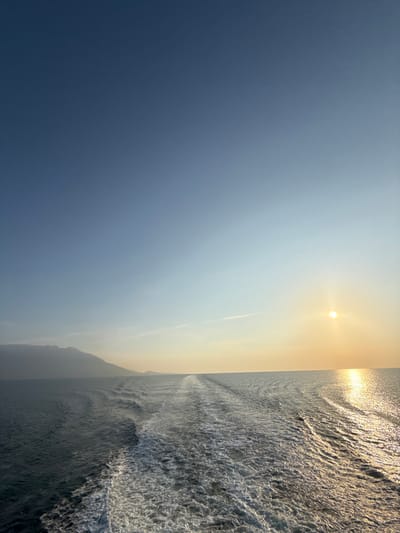The image presents a seascape from the stern of a moving vessel, likely taken near Kato Karioutes, Greece, during either sunrise or sunset. A prominent white foamy wake extends directly from the foreground to the distant horizon, indicating the vessel's forward motion. To the left, a dark, low-lying mountainous landmass is discernible along the horizon. On the right, a bright, circular sun sits low on the horizon, casting a strong golden light path across the water's surface towards the viewer. The sky transitions from a deep blue in the upper left to a lighter, golden-orange hue near the sun, with a faint linear contrail visible in the upper right quadrant. The water is dark blue-green, displaying surface ripples and the agitated foam of the wake. No individuals or specific man-made objects other than the implied vessel creating the wake are present.