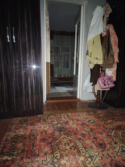 The image depicts an interior scene, likely a hallway or entrance area in a home. On the left, a dark, vertical-grained wardrobe or cabinet dominates the frame. Its surface is reflective, showing bright streaks of light. To the right of the wardrobe is an open doorway leading into another room, which is dimly lit and features sheer curtains.
In the foreground, a patterned rug with red, black, and yellow motifs covers the floor. The floorboards beneath the rug are visible in places and appear to be polished wood.
To the right of the doorway, several garments are hanging on a wooden stand. These include a white garment, a yellowish garment with decorative trim, a brown jacket, and an orange and white patterned garment. A small pink handbag with cartoon characters on it is also hanging from the stand. A plastic bag and a dark garment are also present on the stand.
The lighting in the image is dim, suggesting it might be nighttime or a poorly lit indoor space. The overall impression is one of a lived-in domestic environment. No people are visible in the image. There is no visible text. The location context of Akaltyn, Uzbekistan, suggests a Central Asian setting.