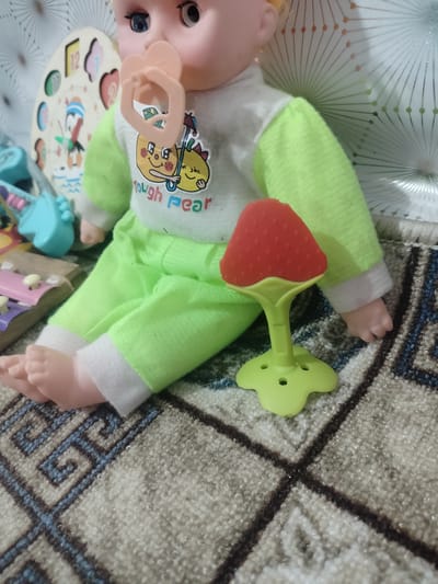 A baby doll is sitting on a patterned rug in what appears to be a home environment. The doll is wearing a white shirt with a green long-sleeved overlay adorned with cartoon characters and the text "Tough Pear," and green pants with white cuffs. A pink pacifier is in the doll's mouth. To the right of the doll is a red, strawberry-shaped teether with a green handle and base. In the background, a wooden clock face with numbers and a penguin illustration is visible, along with other colorful children's toys. The lighting suggests an indoor setting. The image does not contain any information to ascertain its location within the city of Cholpon, Kyrgyzstan.