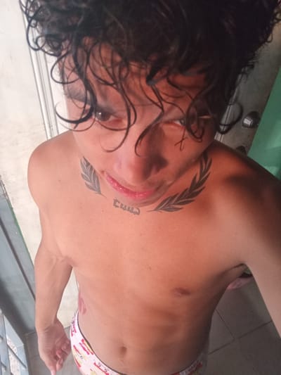 The image shows a shirtless person with dark, curly hair, looking down at the camera. They have tattoos on their neck, one resembling a laurel wreath and another with stylized lettering. The person is wearing patterned underwear. The background appears to be indoors, with a white wall and a green surface visible. The lighting suggests it is daytime. The provided location of La Guardia, Venezuela is not visually verifiable from the image itself.