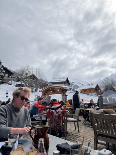 The image captures an outdoor dining terrace in La Clusaz, France, on an overcast day with a uniform layer of grey and white clouds. In the foreground, a Caucasian male with medium-length curly brown hair, wearing dark sunglasses and a grey knitted sweater, is seated at a light-colored table. His right hand is resting on the table. On the table are a clear glass of water, a blue disposable lighter, a dark shaker, slices of bread, a brown ceramic pitcher, and a clear glass bottle.

In the midground, numerous individuals are seated at wooden tables on a timber deck terrace. Patrons are dressed in various winter garments, including a person in a red and white striped jacket with a brown backpack, and another in a red top interacting with someone across a table. A male in a dark vest stands facing away from the camera, positioned among the tables. Ski equipment is visible leaned against a barrier on the left side of the terrace. A red safety net or barrier is present on the snowy slope beyond the terrace.

The background reveals a snow-covered landscape with several chalet-style buildings featuring dark roofs and light-colored walls, nestled among snow-dusted bare trees and evergreens on ascending slopes. A dark SUV is parked on a snowy path behind a chalet on the left. Distant snow-covered mountains are visible under the cloudy sky.