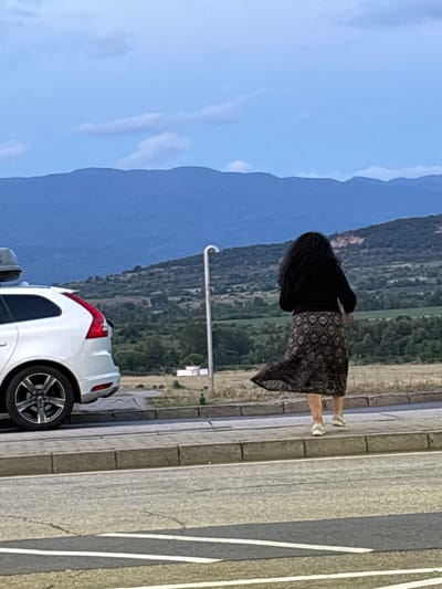 In Kalugerovo, Bulgaria, a woman with long, dark, curly hair walks away from the camera. She is wearing a black sweater and a patterned, flowing skirt. Her legs are bare, and she is wearing white athletic shoes. She is walking on a sidewalk next to a white car. The car is a wagon with a rooftop cargo carrier. The car is parked on the left side of the frame. The background shows a landscape with rolling hills and mountains. The sky is blue with some clouds. There is a street in the foreground with white lines painted on it.