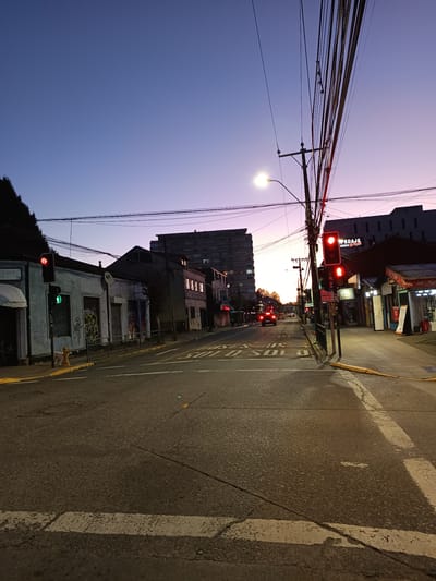 The image captures a street scene in Osorno, Chile, at dusk or dawn. The sky is transitioning from blue to a soft pink.
The street is lined with buildings of varying heights and styles. A mix of residential and commercial properties are visible, including a building with "PEDAJE" visible on the side. On the left, a low building has a green traffic light, while on the right, another traffic light displays red. Power lines crisscross overhead.
The street is paved with asphalt. Markings indicating bus lanes are visible on the road surface. There are few visible people in the frame. A couple of cars are moving further down the street. There is a fire hydrant on the left, near the green light.