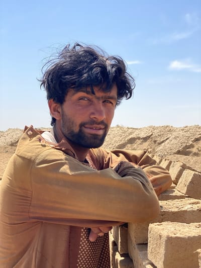 A medium shot depicts a man outdoors. He has dark hair and a beard, and is wearing a brown, patterned shirt with a collar. He is leaning against a low wall of bricks. The background includes a field and a blue sky with a few clouds. The location is Deh-E Bagh, Afghanistan.