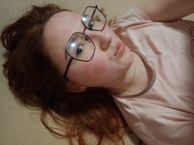A close-up photograph depicts a young individual with reddish-brown hair, lying on a light, neutral-colored surface, positioned diagonally within the frame. The subject wears a light pink t-shirt with a darker pink collar trim and black, hexagonal-framed eyeglasses. A thin silver-colored chain is visible around their neck. The subject's lips are slightly parted and pursed in a pouting expression, with a glossy appearance. Both eyes are visible through the eyeglasses, gazing towards the camera, with light reflections present on the lenses. The cheeks show a flushed complexion. The surrounding environment is an indistinct, uniform light-beige texture. This scene is located in Старый Шагирт, Russia.