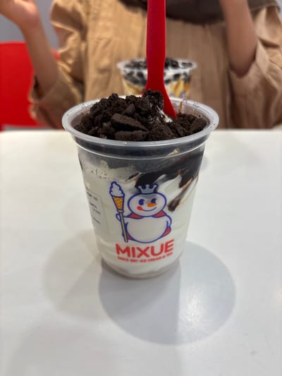 A close-up shot focuses on a clear plastic cup filled with a white creamy dessert topped with crushed dark cookies and drizzled with chocolate sauce. A red plastic spoon with "MIXUE" branding is partially submerged in the dessert. The cup has a snowman logo and "MIXUE SINCE 1997 ICE CREAM & TEA" printed on it. In the background, a person wearing a light brown cardigan is visible, with a blurred red chair on the left. Another blurred cup with toppings is behind the main subject. The scene appears to be indoors, on a white table, possibly at a MIXUE store in Gampongpaya, Indonesia.