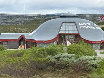 This media file captures an exterior view of the Arctic Center in Norway. The main subject is a unique, dome-shaped building with a grey, rounded roof and red accents. The building features a large, multi-paned window with a red frame on its curved facade. White lettering on the dome reads "ARCTIC CENTER 69° 32'". To the left of the dome, a lower, wooden-clad section of the building with red trim and windows is visible. A flag with red and white stripes and blue elements, likely the Norwegian flag, flies on the right side of the building.
The building is situated in a barren, hilly landscape with sparse vegetation, predominantly low-lying green bushes and some scrub. In the background, the terrain appears rocky and desolate, with patches of snow visible on distant hills, suggesting a cool climate. The sky is overcast with grey clouds, indicating a cloudy or possibly rainy day. The time of day is unclear, but the lighting suggests it is daytime. There are no people visible in the image.
Notable details include the distinctive architecture of the Arctic Center, designed to withstand harsh arctic conditions. The contrast between the modern, rounded structure and the rugged natural environment is striking. The presence of the Norwegian flag provides a clear location cue. The overall scene conveys a sense of remoteness and the challenging conditions of the Arctic region.