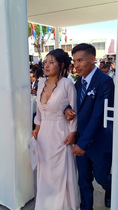 The image shows a couple at an event in Cerro Colorado, Peru. The woman is wearing a light-colored, long-sleeved dress, while the man is in a dark blue suit with a patterned tie and a white flower boutonniere. They are standing under a white tent-like structure. In the background are other people and buildings, with colorful flags visible. The woman is holding a document with a crest on it.
