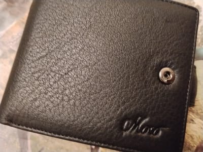 The image shows a close-up, overhead view of a dark brown, textured leather bi-fold wallet. The wallet is lying flat. A silver snap closure with an embossed logo is visible on the right side. Embossed in the bottom right corner of the wallet is a cursive logo that appears to read "Noco". The background is a light-colored, patterned surface. There are no people, actions, or interactions depicted. The location is unknown, and no specific details about Iziaslav, Ukraine are visible.