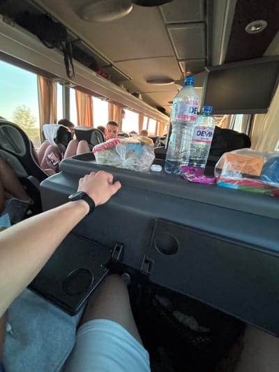 A bus interior is shown, with a person sitting in a seat and another hand reaching to adjust the tray table. Two bottles of water labeled "DEVIN" and some snacks sit on the tray table. People are seated in the rows of seats further down the bus. The image appears to be taken during a trip, though the location in Bošnjaci, Croatia, is not visually verifiable from the photo.