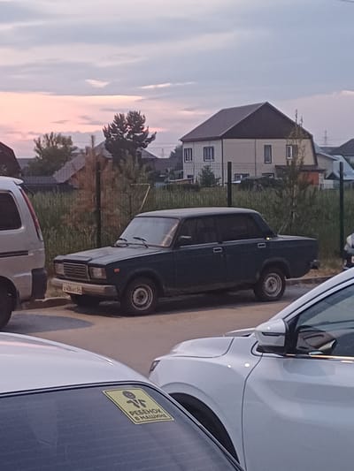 This is a slightly tilted, outdoor, eye-level, close-up photograph taken in Elitnyi, Russia. The image features several vehicles parked close together on a paved surface, likely a street or parking lot, with residential buildings and greenery in the background. A dark-colored sedan is prominent in the center, with its front and rear visible. To the right, a portion of a white car is visible, with a yellow sticker on its rear window. The sticker has text in Russian, reading "РЕБЕНОК В МАШИНЕ," which translates to "Child in the car," and a pictogram of a child. The sky in the background appears overcast with hints of pink and orange, suggesting it is either dawn or dusk. The overall mood is calm and ordinary, depicting a typical day in a residential area.