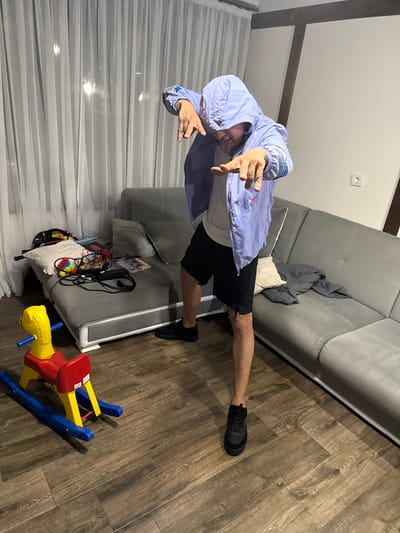 An indoor scene in Hristo Danovo, Bulgaria, depicts a male individual posing in a residential room. The person, positioned slightly right of center, wears a light blue hooded windbreaker over a white t-shirt, black shorts, and black athletic shoes. The jacket displays an "adidas" logo on the left chest. The individual's hood is up, covering the head, and a portion of the face shows an open mouth. Both arms are extended forward and slightly outward, with hands open, fingers splayed in a claw-like gesture.
The room features dark wood laminate flooring. Along the back wall and extending to the right is a large L-shaped grey faux leather sectional sofa with white trim at its base. The left section of the sofa is cluttered with various items: a black bag, two pillows (one white, one light grey), a tennis racket handle, a colorful child's toy, a video game case, and an assortment of tangled black electronic cables and devices, including a black console. On the right section of the sofa, another light grey pillow and a dark grey blanket or piece of clothing are present.
The entire back wall is covered by sheer white curtains, behind which diffused light sources are visible. The right wall is light-colored, featuring a white electrical outlet with two sockets. A dark wooden beam structure is visible intersecting the upper right wall and ceiling. In the lower left foreground, a yellow and red child's rocking horse with blue rockers rests on the floor.
