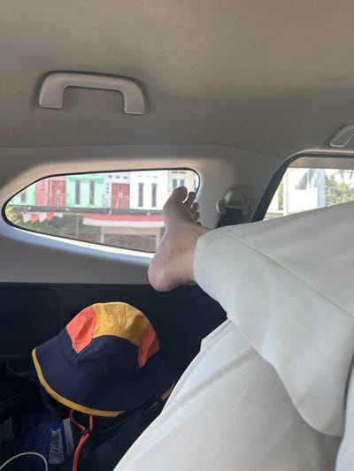 The image shows a person's bare foot and lower leg, clad in white pants, resting against the interior of a vehicle. The foot is positioned near a car window, through which a view of buildings can be seen. The buildings are colorful, with sections painted red, white, and green, suggesting a tropical or subtropical location. The sunlight filtering through the window indicates it is daytime. A multi-colored bucket hat, with navy blue, yellow, and orange panels, lies on the floor of the vehicle next to the person's leg. The interior of the car is dimly lit, with a grab handle visible on the ceiling. The scene appears casual and relaxed, possibly during a road trip. The overall impression is one of comfort and leisure.