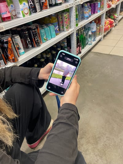 A person is holding a smartphone with a picture of a young man in a striped shirt and black pants standing on a sidewalk. The phone is in the foreground, and the background shows shelves stocked with various drinks, including energy drinks and bottled water, in what appears to be a convenience store or supermarket in Alyumino, Russia. The person's hands are visible holding the phone, and they are wearing a black jacket and black pants. The overall lighting is artificial, typical of an indoor retail environment.