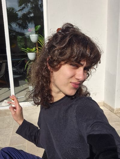An individual is depicted in a close-up selfie format, positioned outdoors on a tiled patio or terrace in what appears to be a residential setting within Châtelaillon-Plage, France. The subject is a person with medium-brown, curly hair, partially pulled up in a bun secured with a small, orange hair claw clip, while bangs frame the forehead. The individual wears a dark navy or black long-sleeved crew-neck top and dark blue pleated trousers, visible at the lower left. The left hand is raised, with fingers splayed, holding a small, cylindrical light-colored object between the thumb and index finger. The subject looks directly at the camera with a slight smirk.
Behind the individual, a white wall, strongly illuminated by sunlight, occupies the right side of the frame, indicating direct sun exposure. To the left, a large glass sliding door or window with a white frame reflects palm trees and an overcast sky. A multi-tiered stand holding two white potted plants is also visible in the reflection of the glass. The ground surface is paved with light-colored square tiles. Shadows are visible, consistent with bright daylight.