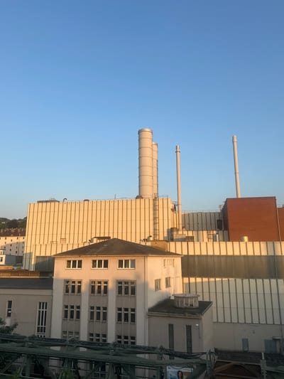 The image shows a panoramic view of an industrial complex in Wuppertal, Germany, under a clear blue sky. The main structure is a large, light-colored building with a corrugated facade, possibly a factory or power plant, featuring several tall, cylindrical smokestacks. To the left of this large building, a smaller, multi-story building with numerous windows is visible. In the foreground, a dark, metallic structure, possibly a bridge or walkway, stretches across the bottom of the frame. The overall scene conveys a sense of industrial activity within an urban setting.