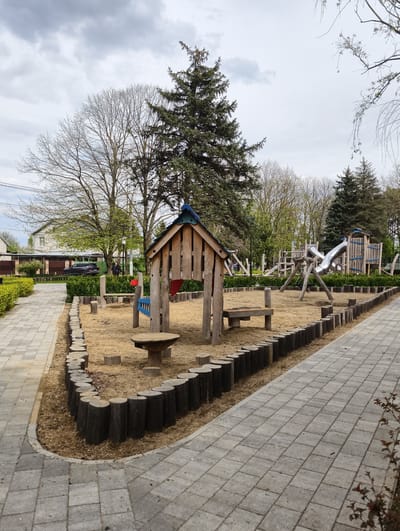A playground is present in Kolosisty, Russia, located outdoors on a cloudy day. It contains a wooden playhouse with a blue roof, a table, benches, and metal slides, all surrounded by sand and logs. A paved walkway borders the playground. Trees and a building are visible in the background. Two people are visible in the distance. A dark-colored SUV is parked nearby.