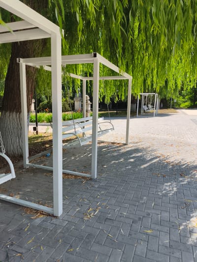 A public park scene in Ocnița, Moldova, features a white metal swing set with a bench swing, situated under the drooping branches of a large willow tree. The ground is paved with gray interlocking bricks, scattered with fallen yellow and brown leaves. In the background, another white metal swing set is visible, along with a statue. The area appears to be a recreational space within the city, designed for public use.