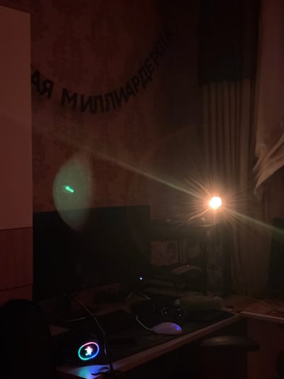 In a dimly lit room, likely a bedroom or study, a banner with Russian lettering is hung on a patterned wallpapered wall. The banner appears to read "Я МИЛЛИАРДЕРША" which translates to "I am a billionaire". A bright light source, possibly a lamp or flash, is positioned in the middle right of the frame, emitting strong rays that create a lens flare effect. In the foreground, a computer monitor is visible, along with a keyboard and mouse. One mouse is illuminated with colorful LED lights, featuring a glowing animal emblem. A green light artifact is present on the left side of the monitor. Curtains are drawn on the right side of the room, and a portion of a beige curtain is visible at the top right. The overall scene suggests a personal space, possibly decorated for a celebration or a personal declaration. The image does not provide any specific information about the city of Radkevshchina, Russia.