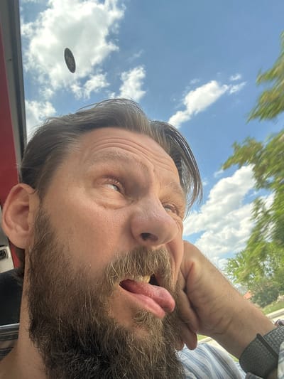 The original image is a close-up, wide-angle selfie taken from a low perspective, looking upwards. The subject is a man with a beard and shoulder-length brown hair, wearing a blue and white striped shirt. He is making a comical facial expression, with his eyes rolled back and his tongue sticking out. His left hand is resting on his cheek. In the background, there is a bright blue sky with scattered white clouds, and blurry green trees are visible to the right. A dark, circular object, possibly a coin, is suspended in the air in the upper left portion of the image. The image appears to be taken inside a vehicle, indicated by the red frame on the left and the reflection on the windshield. The scene is set in the city of Çavdır, Turkey, during the daytime.