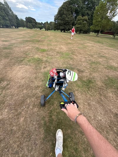 This image captures a golf course scene in Brandesburton, United Kingdom, during daylight hours under a partly cloudy sky. The perspective is first-person, showing the subject's right hand gripping the black handle of a blue two-wheeled golf push trolley. The subject's hand displays rings on the index and middle fingers and a metallic chain bracelet on the wrist. The lower part of the subject's right leg, clad in a light-colored athletic shoe and white socks, is visible.
The blue golf trolley holds a black golf bag containing multiple golf clubs, identifiable by various headcovers, including a red one with a white cross and a white one with green trim. The fairway is characterized by a mix of dry, brown grass and greener patches. In the mid-distance, approximately 50 to 70 meters away, another individual, dressed in white shorts and a red and white short-sleeved top, is walking away from the camera, carrying a white golf stand bag. The golf course is bordered by numerous mature trees. A small, reddish building is visible among the trees on the right side of the frame, and a white golf flagstick is discernible further down the fairway in the distance.