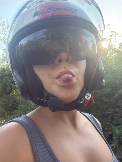 A close-up, self-portrait style image shows a woman wearing a motorcycle helmet with a reflective visor. Her mouth is slightly open, with her tongue sticking out. She is wearing a grey tank top. The background is blurred, showing green foliage and a bright sunburst in the upper right corner, suggesting an outdoor setting, possibly in Buljarica, Montenegro, during daylight.