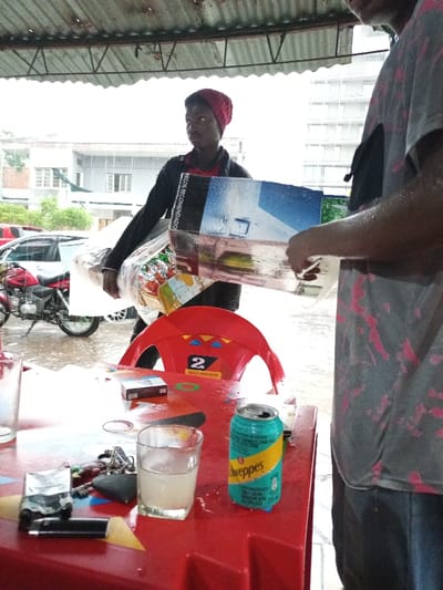 This image captures a rainy daytime scene in what appears to be a sheltered outdoor or semi-outdoor area, possibly a street vendor stall or cafe, consistent with the context of Maputo, Mozambique. Heavy rain is visible as streaks against the bright, overcast background.

Two men are present. On the left, a man in a red beanie and dark jacket carries a large clear plastic bag filled with goods and a rectangular object that displays an image of a modern house and bears the text "PREÇOS RECOMENDADOS" (Recommended Prices). He looks towards the left with a neutral expression. To the right, another person is partially visible, wearing a grey t-shirt with a pink pattern, seemingly holding or interacting with the rectangular object.

In the foreground, a vibrant red plastic table holds various items: an open turquoise can of "Schweppes," a clear glass containing a light-colored liquid, a cigarette pack, a black lighter, and a set of keys with a colorful keychain. A red plastic chair, marked with the number "2" and "MAC-MAHON," sits near the table. Beyond the covered area, a red motorcycle and a car are parked, while several buildings, including a multi-story structure and a residential house, are visible in the rainy background. The corrugated metal roof overhead offers shelter from the downpour.