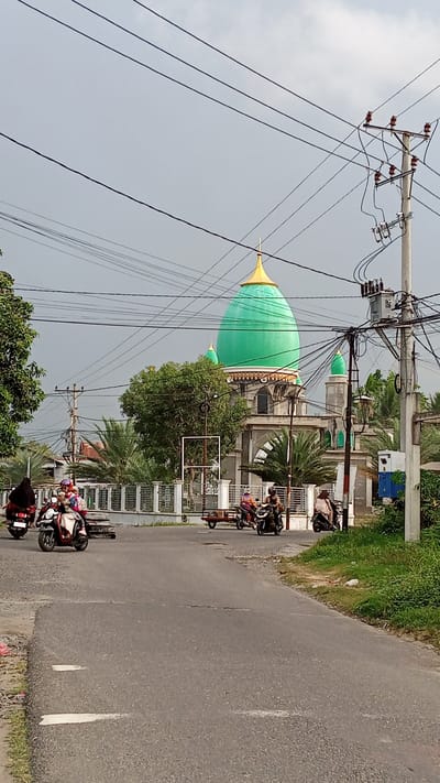 The image depicts a street scene in Geulanggang Teungoh, Indonesia, featuring a prominent mosque with a large green dome topped with a golden spire. Several people are seen riding motorcycles on the paved road in front of the mosque. The mosque has grey stone walls and arched windows, with smaller green domes on either side of the main dome. Power lines crisscross the sky above the scene, supported by utility poles. Trees and palm trees are visible around the mosque and along the roadside. The road appears to be in a somewhat rural or suburban area, with a white fence bordering the mosque property. The sky is overcast.