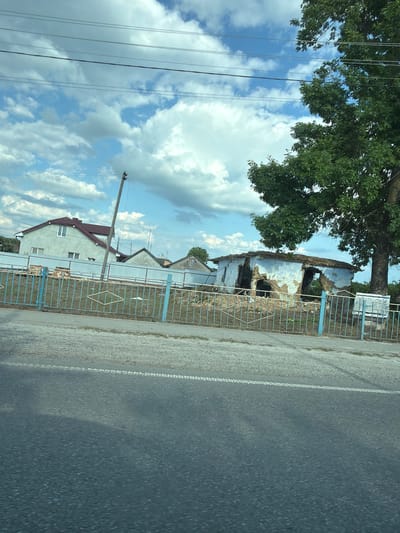 This media file depicts a roadside scene in Teofipilka, Ukraine, likely during the daytime under a cloudy sky. The image is oriented vertically.
The foreground is dominated by a paved road with a white dashed line marking its edge. To the left of the road, there is a gravel shoulder and then a blue metal fence. Beyond the fence, several structures are visible. A large, mature tree with lush green foliage stands prominently on the left, its branches extending over the fence and a damaged building. This building appears to be an old, partially collapsed structure with exposed walls and a gaping roof, suggesting it may be abandoned or has suffered damage.
Further down the fence line, there are houses with visible roofs and windows, one house being particularly prominent with a red-tiled roof and light-colored walls. Utility poles with overhead wires are also present, indicating the presence of infrastructure. The sky is a mix of blue and white clouds, suggesting a typical summer day. The overall impression is one of a rural or semi-rural setting with a contrast between newer residential buildings and older, decaying structures. There is no discernible activity or people in the image. No visible text is present.