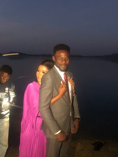 A man in a gray suit and red tie stands next to a woman wearing a magenta dress with her arm around him. Both smile warmly. In the foreground, a man in a dark shirt looks down with a light shining on his hands. The background shows a dark body of water meeting a dark sky. A small patch of vegetation is visible on the right side of the image.