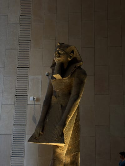 This media file showcases a large, ancient stone sarcophagus lying horizontally against a stone-tiled wall. The sarcophagus depicts a human figure, likely a pharaoh, in a stylized, elongated form. The figure has a distinctly Egyptian headdress and facial features. The stone is weathered, showing intricate carvings and the passage of time.

The sarcophagus is illuminated by warm, directional lighting, casting dramatic shadows that highlight the texture and form of the sculpture. The wall behind it is composed of large, rectangular beige tiles, creating a modern and museum-like setting. Below the sarcophagus, a series of rectangular grates or vents are embedded in the wall.

Visible in the upper right corner of the image, a person is partially visible, leaning forward and looking at the sarcophagus, suggesting the setting is a museum or historical exhibition. There is no visible text in the media file. The lighting suggests an indoor environment, possibly with controlled lighting for display purposes, rather than natural daylight. The overall impression is one of historical grandeur and preservation within a contemporary exhibition space.