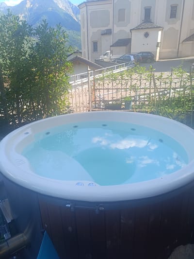 The image shows an outdoor hot tub with clear blue water, reflecting a bright sky with scattered clouds. The hot tub is round with a white rim and a wooden exterior. In the background, there is a white-gated fence and lush green trees. Beyond the fence, a building with an architectural design typical of alpine regions is visible, with a sloping roof and stone accents. The building appears to be a church or historical structure. Further in the distance, imposing mountains with rocky peaks and forested slopes rise under a clear blue sky. Several cars are parked on a paved area in front of the building. The scene suggests a peaceful setting in a mountainous region. The location is identified as Serravalle, Switzerland.