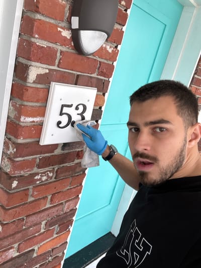 This image depicts a young man engaged in cleaning a house number plaque on the exterior of a building, likely a residence in Hampton, United States of America. The photograph is taken from a low, tilted angle, looking upwards.
The main subject is a man with short dark hair, a beard, and dark eyes, who appears to be in his 20s or 30s. He is wearing a black t-shirt with a white, stylized graphic design on the front. On his left wrist, he wears a dark-strapped watch, and his right hand is covered by a light blue latex or nitrile glove, holding a light-colored cloth or rag. He is looking directly towards the camera with a focused expression.
The activity taking place is the cleaning of a white rectangular house number plaque, which clearly displays the black numbers "53". The man's gloved hand and cloth are positioned over these numbers. The plaque is mounted on a red brick wall, characterized by visible white mortar and some areas of wear or efflorescence. To the left of the plaque, a dark grey outdoor wall light with a textured lens is partially visible. In the background, above the brick wall, a vibrant turquoise-colored door or door frame with white trim provides a contrasting element.
The scene is set outdoors during what appears to be clear daytime conditions, given the bright and even lighting. The visual cues suggest a routine maintenance task.