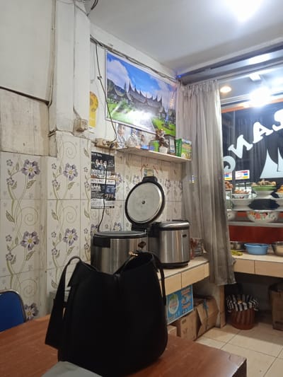 The scene is an interior shot of a restaurant or shop in Kecamatan Kartasura, Indonesia. The image appears to be taken near a counter, and there is a white tiled wall covered in a floral pattern to the left, and a window to the right. There is a black purse on the counter, in front of an open rice cooker and an electric rice cooker. On the wall to the left of the rice cookers there is a poster. To the right of the rice cookers there is a window looking out onto a street, which has a red and white sign with the word “MAS” on it. The window is in a room with a white tiled wall and a counter made of light-colored wood. The window is partially obscured by a white curtain, with brown and blue decorations above it. The room has a white ceiling with a single light fixture.