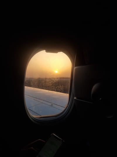 The image provides a perspective from inside an airplane, looking out through an oval window during either sunrise or sunset. A brilliant, low-hanging sun dominates the upper left portion of the window, casting a warm, golden glow across the sky and atmosphere. Below the sun, a hazy landscape, possibly featuring agricultural patterns or distant urban elements, is visible, albeit indistinctly. The metallic surface of an airplane wing extends into the right side of the window's frame.

The airplane's interior is significantly underexposed, appearing almost entirely black, which accentuates the bright external view and the golden light. Only the silhouette of the window frame and overhead cabin structures are faintly discernible. On the far right, a hand is visible, holding a smartphone that is directed towards the window, suggesting the person is taking a photograph or video of the scenic view.

The time of day is clearly twilight, characterized by the intense, low sun and warm light. The weather appears clear, with no clouds obstructing the sun. Faint white text, "S-2B 1/432B", is visible on the lower left edge of the airplane window frame. While the provided location context is Clichy, France, the image itself depicts an aerial view from an aircraft in flight, offering no specific geographical landmarks identifiable as Clichy.