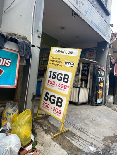 A yellow sign with the words "Zavin Com" and "im3" on it is leaning against a building. It is advertising data plans for a mobile phone provider. The sign lists data plans for 15 GB, 9 GB, and 5 GB. There are a few garbage bags in the foreground, and a partially obscured sign in the background. The location is in the city of Kecamatan Cimahi Selatan, Indonesia at latitude -6.932 and longitude 107.564. The ground is concrete and the sign is standing on a metal frame. A person is partially visible in the background, standing in front of a counter.